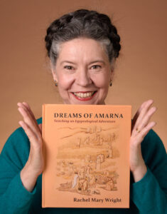 Me and My Book A white woman with dark hair, greying at the temples is - let's be honest - grinning at the camera. Between the flat palms of her hands she is holding up a copy of the book, "Dreams of Amarna - Stitching an Egyptological Adventure".
